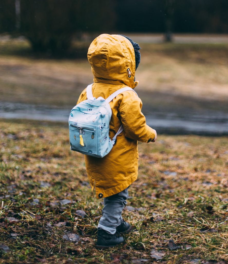 Back of small child wearing a yellow hooded jacket and a light blue backpack walking on grass towards a path