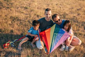 Two fathers holding a rainbow kite with their young children sitting in a field